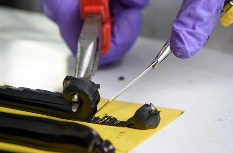 Schwarzer Klebstoff auf gelber Platte, eine Hand entfernt den Klebstoff mit einer Zange