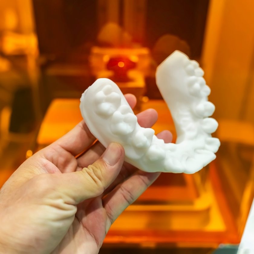 A hand holds a 3D-printed row of teeth against an orange background